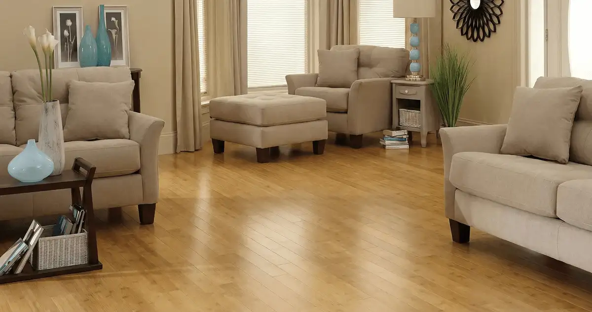 A living room with professionally installed bamboo flooring, beige sofas, a matching ottoman, a side table with a blue lamp, vases, framed pictures, and large windows with blinds and curtains in Milwaukee, WI